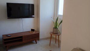 Un salon doté d’un téléviseur à écran plat fixé au mur, d’un meuble de rangement en bois, d’une petite table ronde agrémentée d’une plante en pot et d’une fenêtre laissant entrer la lumière naturelle.