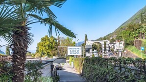 A residential area named 'RESIDENZA LAGO DI LUGANO' with a paved road, a palm tree, and a mountain in the background. - Bissone   Campione Lake Pool & Relax (Bissone)
