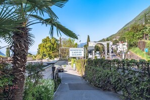 Ein Wohngebiet namens 'RESIDENZA LAGO DI LUGANO' mit einer asphaltierten Straße, einer Palme und einem Berg im Hintergrund.