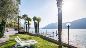 A lakeside setting with a lounge chair, palm trees, a lamp post, and a building with a tiled roof. - Bissone   Campione Lake Pool & Relax (Bissone)