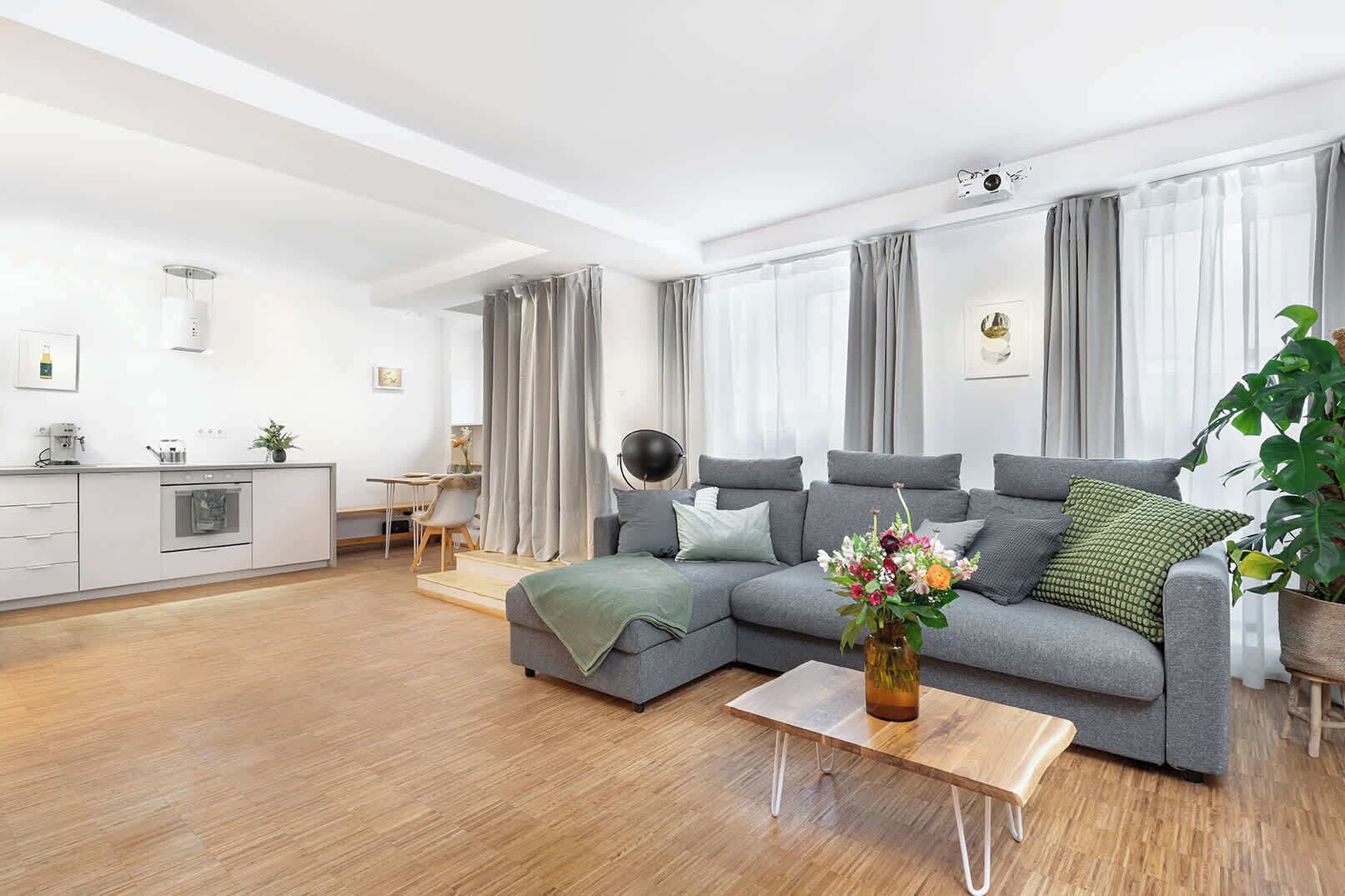 A modern living room with a grey sofa, a coffee table, a dining area with a table and chairs, and a kitchen in the background.