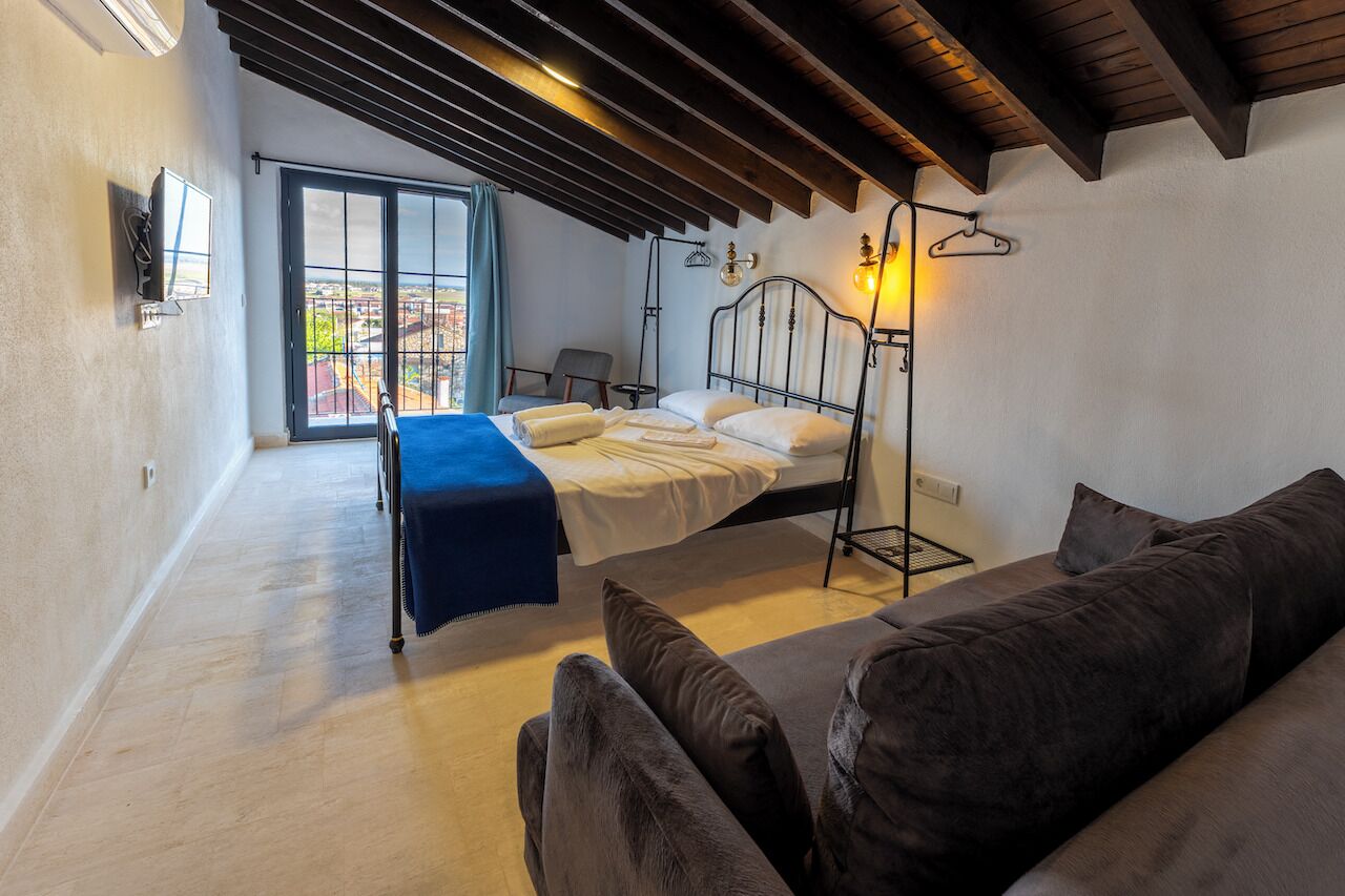 Un dormitorio con cama, manta azul, lámpara colgante, silla y vista por la ventana.