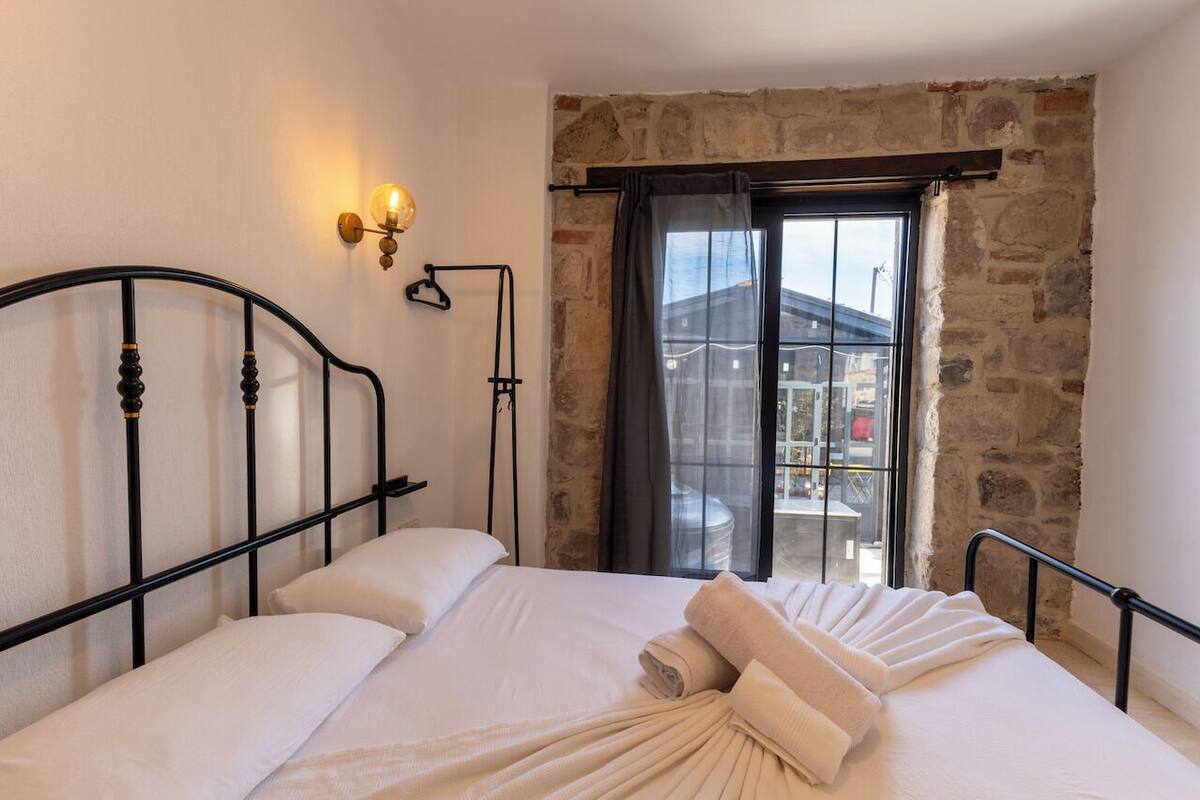 Un dormitorio con pared de piedra, cama con ropa blanca, cabecero de metal negro y ventana con vista al exterior.