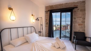 Un lit bien fait, avec du linge de lit blanc, une tête de lit en métal noir et une vue sur la ville par une grande fenêtre ornée d’un rideau noir.
