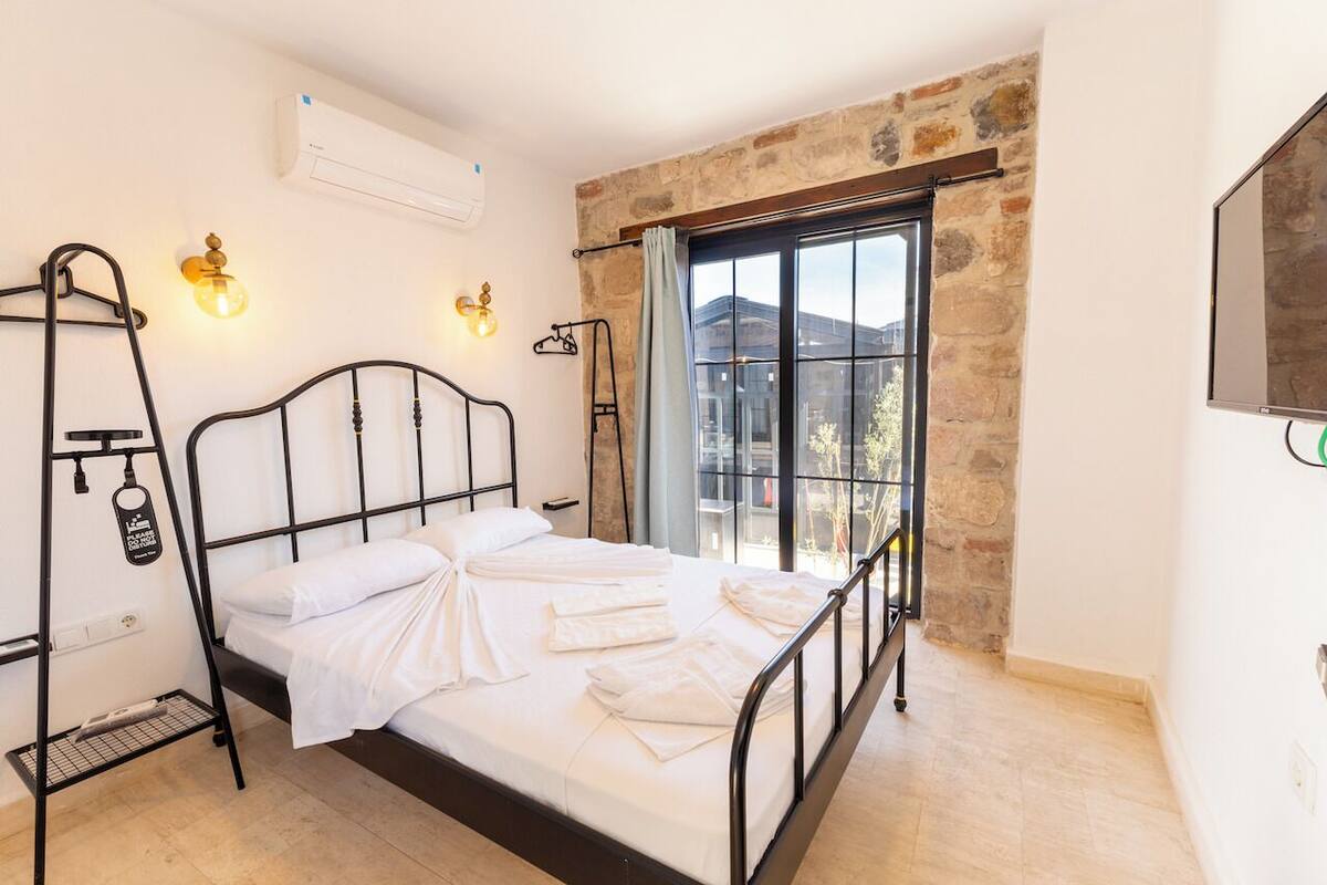 Una cama con ropa de cama blanca, estructura de metal negro, aire acondicionado instalado en la pared, una ventana con cortina y un televisor en la pared.