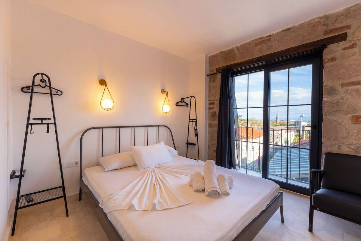 Un dormitorio con una cama, un perchero metálico negro, una silla negra y vistas de la ciudad a través de grandes ventanales.