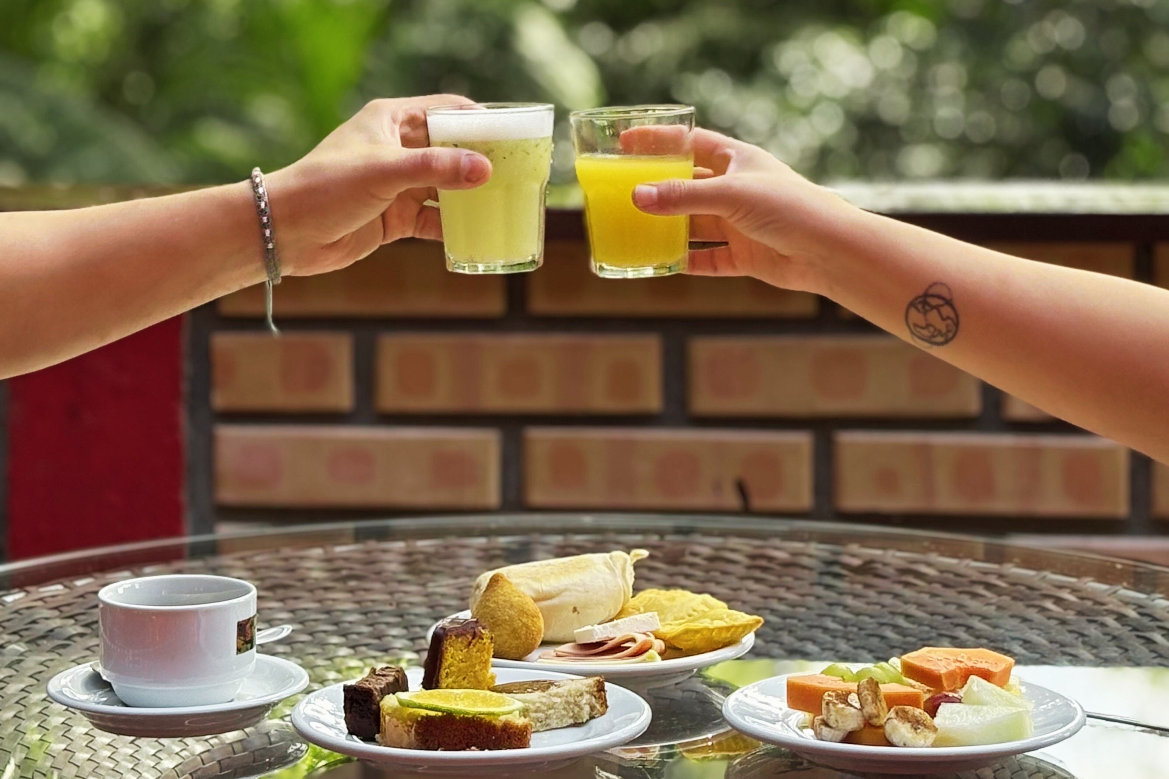 Dos personas brindando con bebidas, una mesa con comida y una taza, y un muro de ladrillo al fondo.