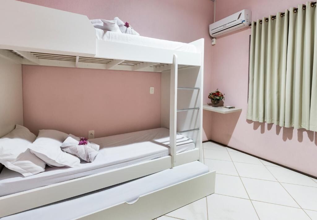 Dormitorio con literas, colchón blanco, almohadas y una pequeña estantería con flores.