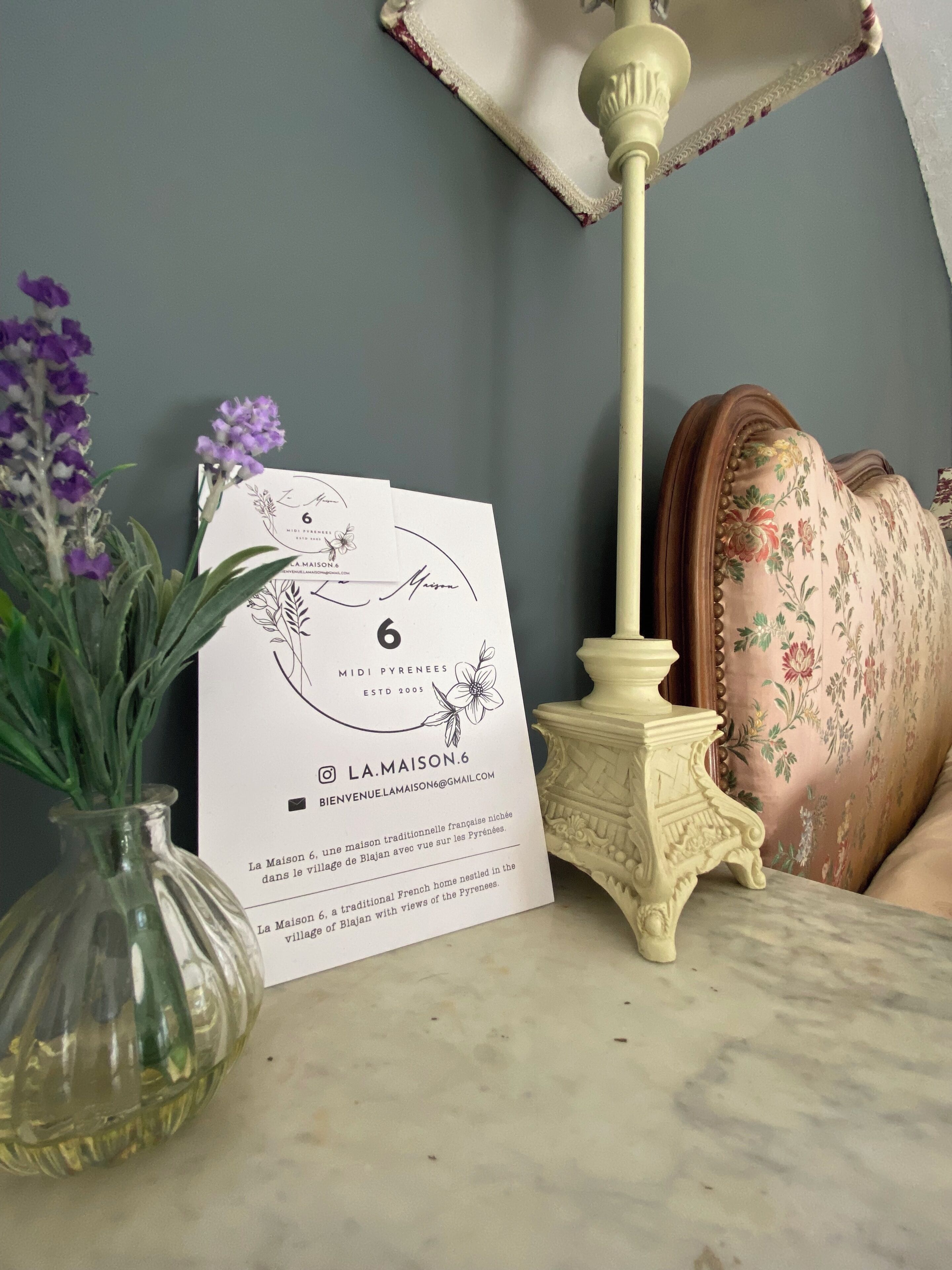 Una tarjeta de bienvenida enmarcada, un florero con flores moradas y una lámpara sobre una mesa.