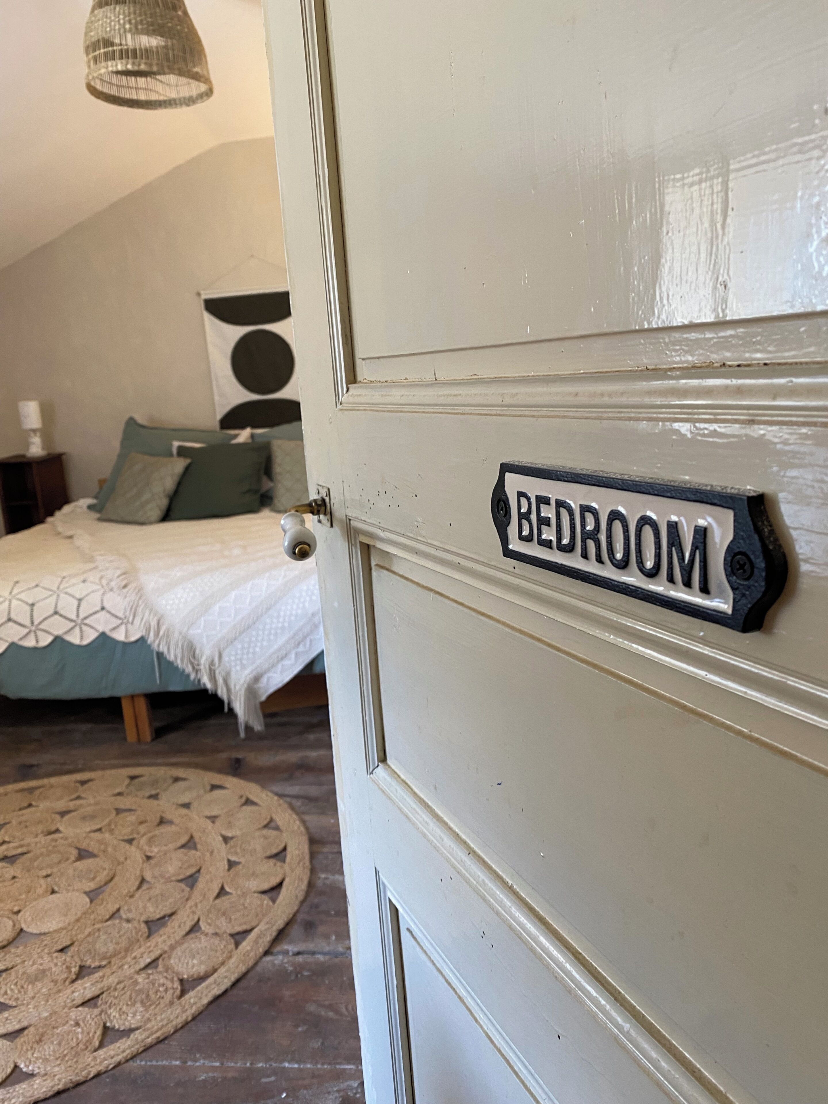 Quarto com porta identificada como 'BEDROOM', cama com colcha verde, luminária pendente e tapete tecido no chão.