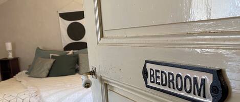 Quarto com porta identificada como 'BEDROOM', cama com colcha verde, luminária pendente e tapete tecido no chão.