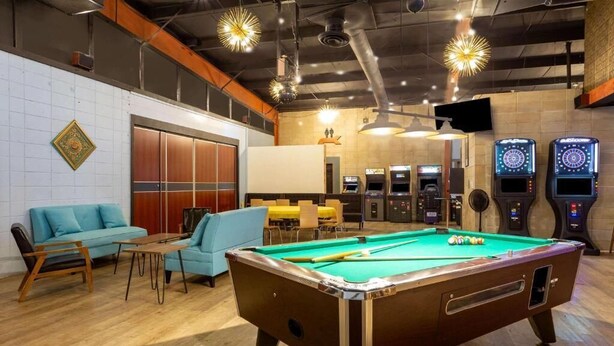 A pool table with surrounding seating, a dartboard, and arcade machines in a room with wooden floors and exposed ceilings.
