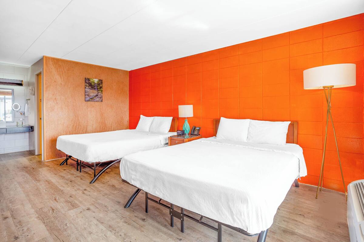 A hotel room with two beds, a wooden floor, an orange accent wall, a bedside table with a lamp, and a painting.