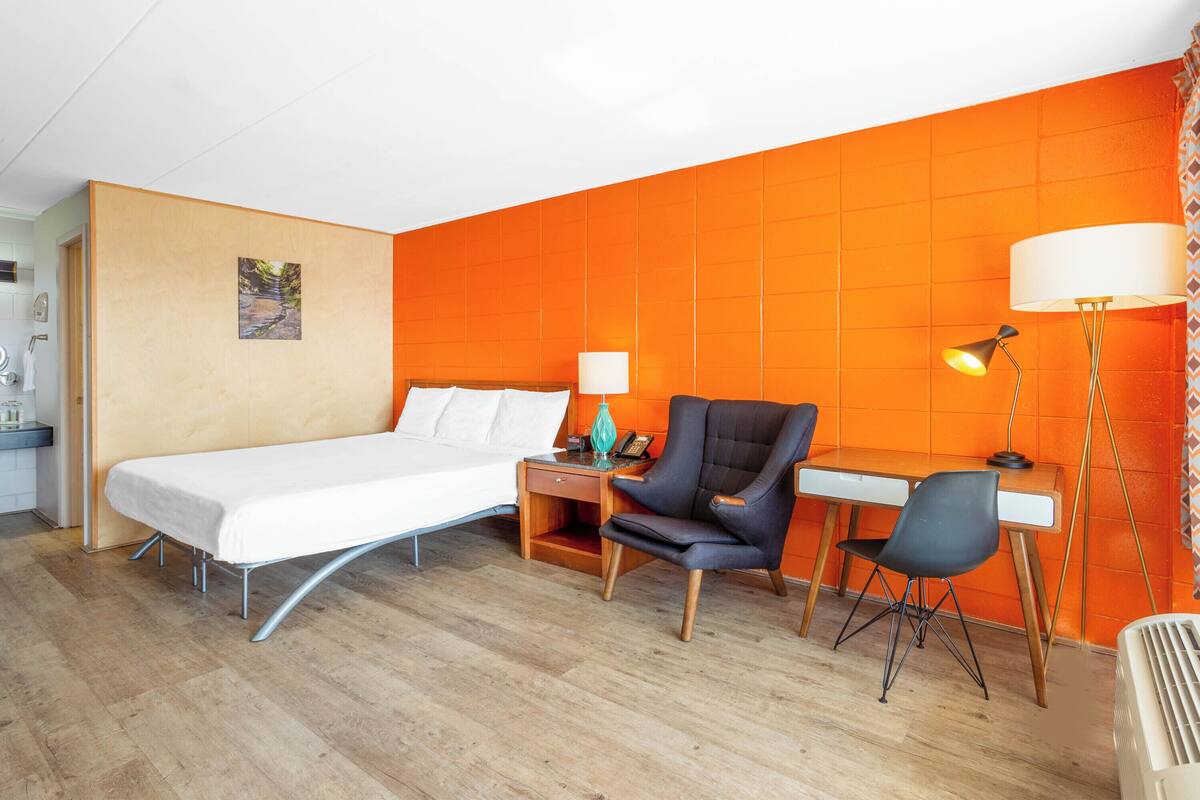 A hotel room with a bed, desk, chair, and a lamp.