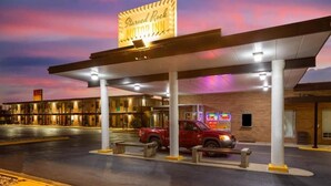 A motel with a neon sign, a covered parking area, and a red vehicle parked inside.