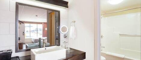 Un baño con lavabo blanco, encimera oscura, espejo y zona de ducha.