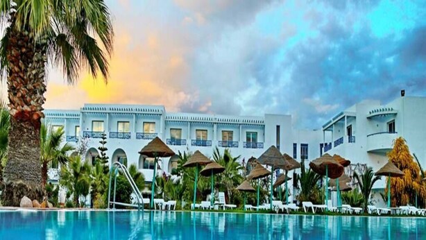 Ein Hotelpool mit Liegestühlen und Sonnenschirmen, umgeben von Palmen und einem Gebäude mit zahlreichen Fenstern.