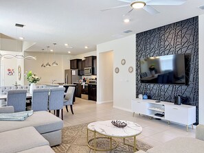 A modern living room with a dining area, kitchen, and a TV mounted on a geometric-patterned wall. - Exquisite 7 beds Pool Home minutes from Disney World! (Davenport)