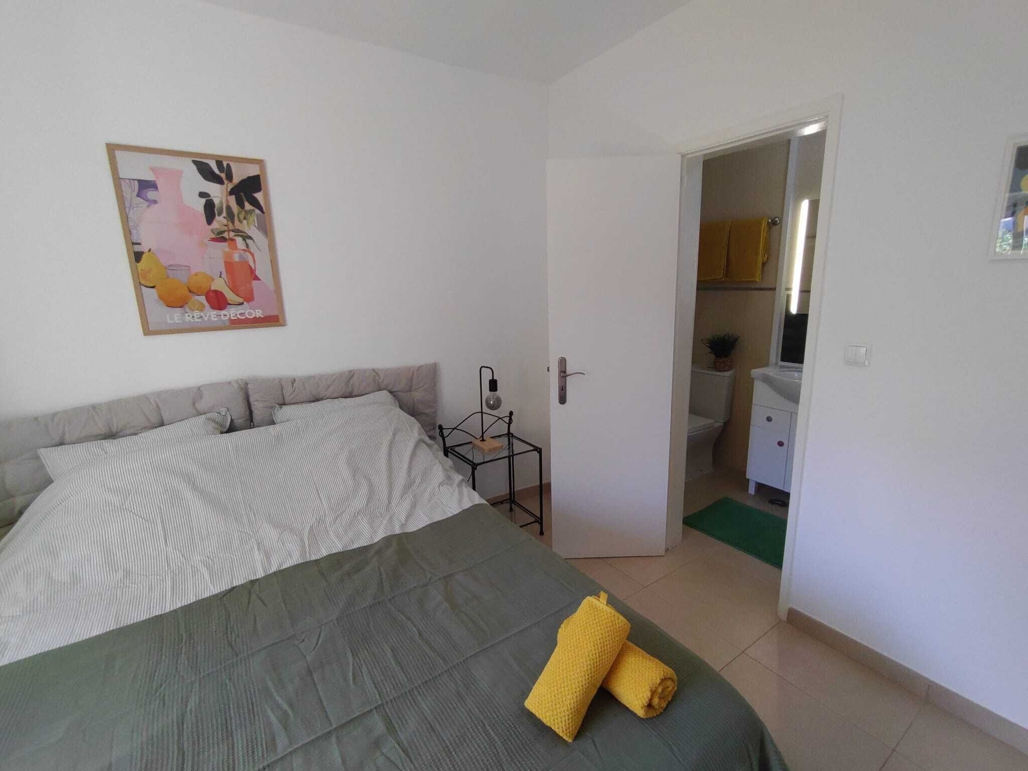 A bedroom with a bed, a bedside table, a nightstand, a door to a bathroom, and a poster on the wall.