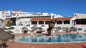 A swimming pool with a unique sculpture, white buildings with red roofs, and a clear blue sky.