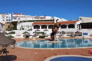 A swimming pool with a unique sculpture, white buildings with red roofs, and a clear blue sky.