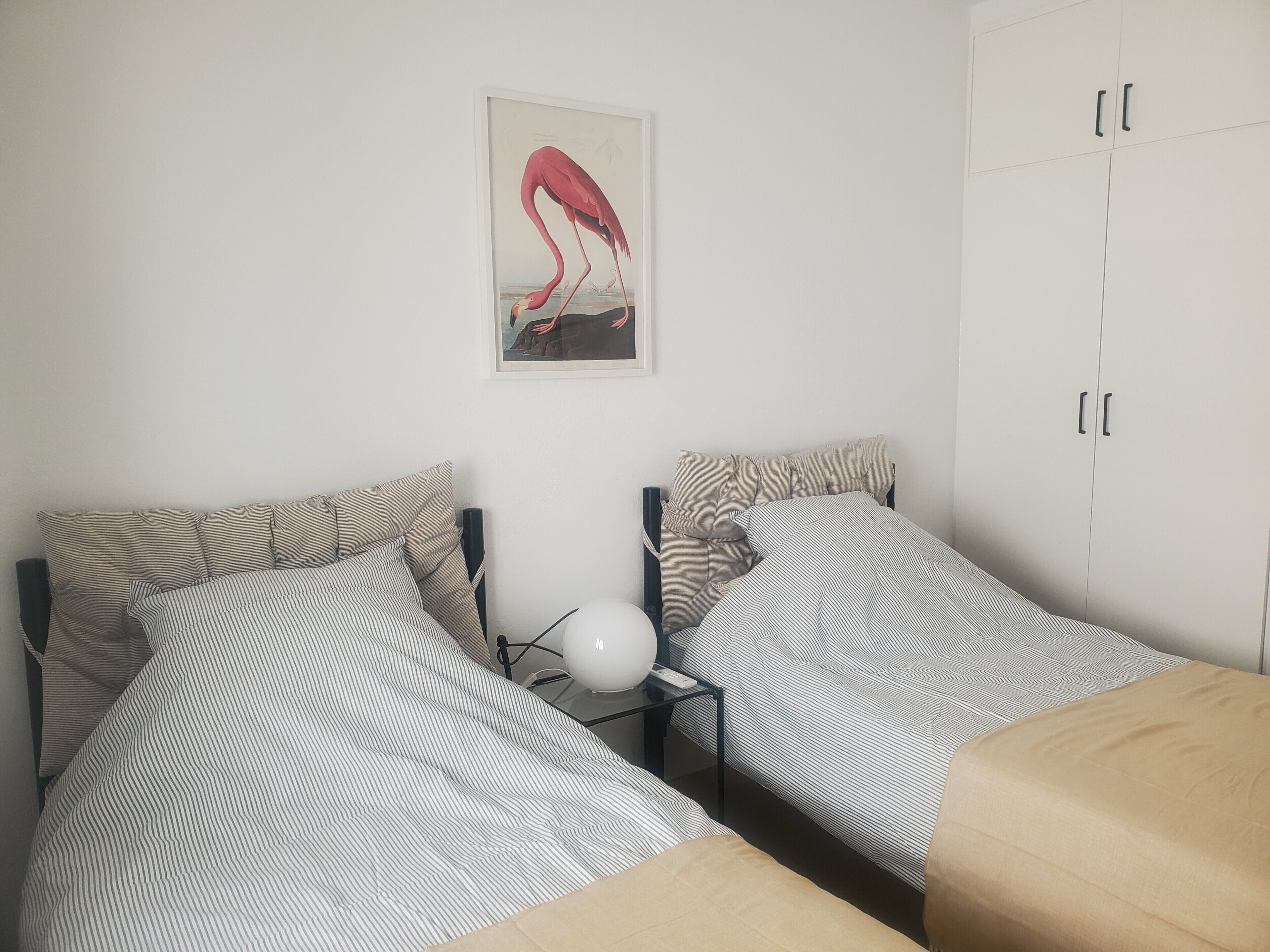 Een slaapkamer met twee bedden, een nachtkastje, een lamp, een kledingkast en een lijst met een afbeelding aan de muur.