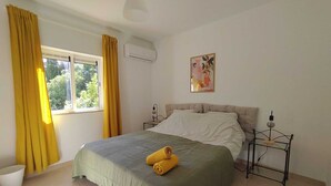 A bedroom with a bed, curtains, a window, an air conditioner, a bedside table, a lamp, and a painting on the wall.