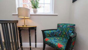 Una habitación de esquina con una silla negra, una mesita con una lámpara y una ventana con una planta.