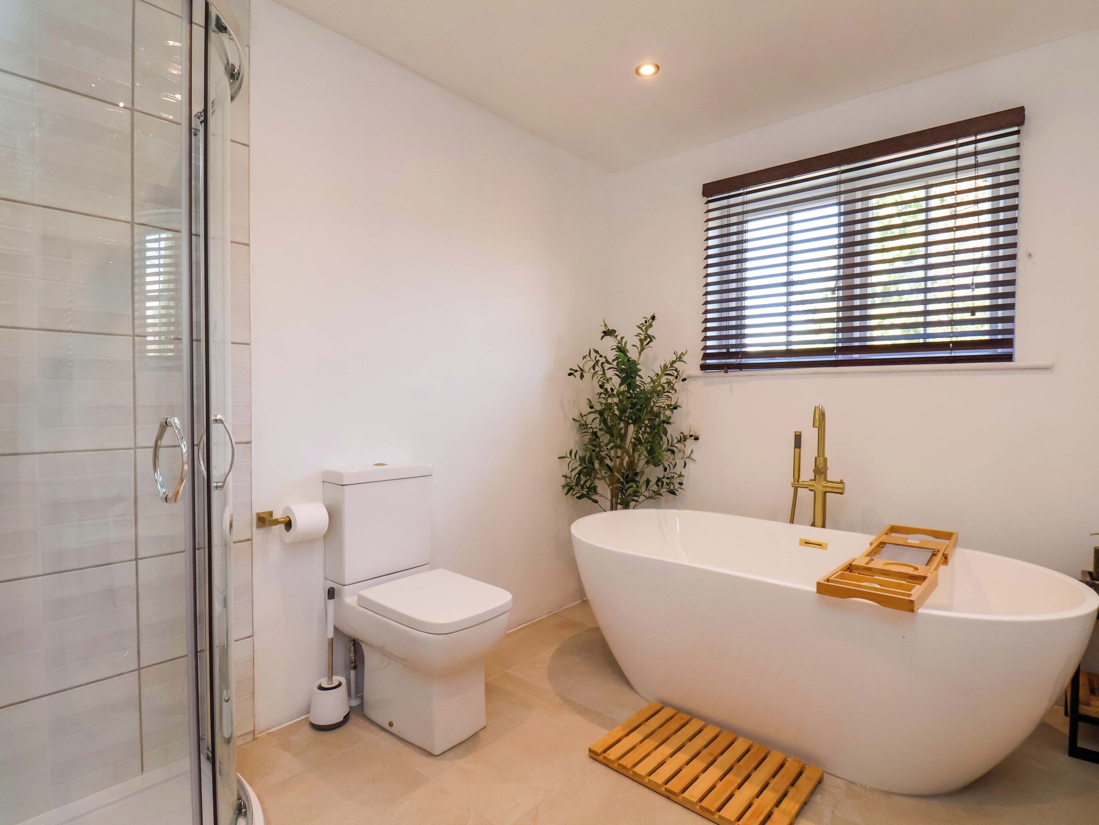 A modern bathroom with a glass shower enclosure, a toilet, a freestanding bathtub, and a window with blinds.
