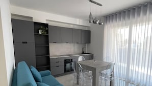 Una sala de estar moderna con un sofá, un comedor con mesa y sillas, una cocina con armarios y electrodomésticos, y ventanas amplias con cortinas.