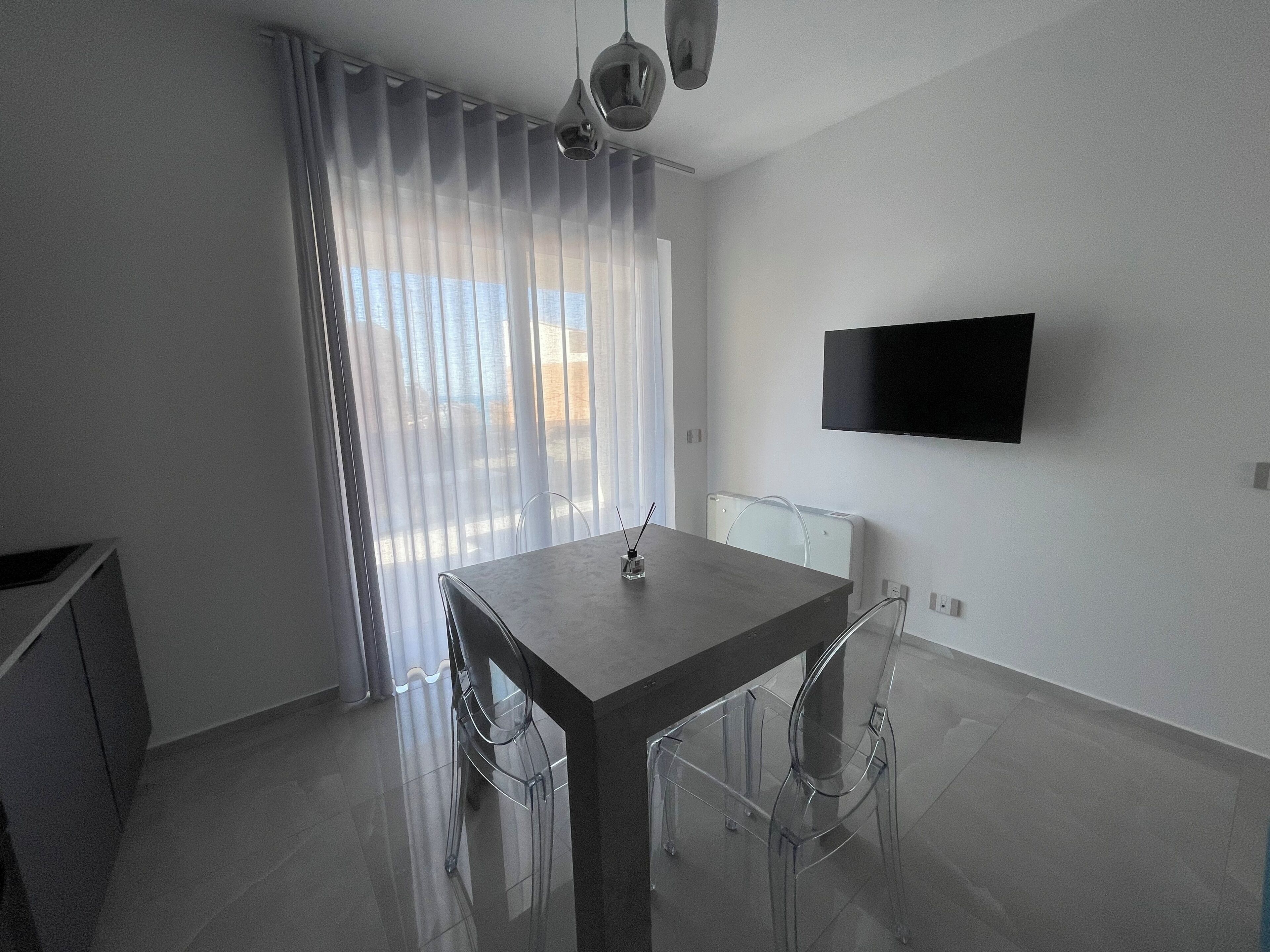 Un espace de repas moderne, avec une table et des chaises, un téléviseur fixé au mur et une fenêtre agrémentée de rideaux légers.