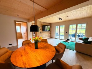 Een ruime woonkamer met een ronde houten tafel, comfortabele zithoek, een aan de muur bevestigde televisie en een glazen schuifdeur die uitkomt op een terras.