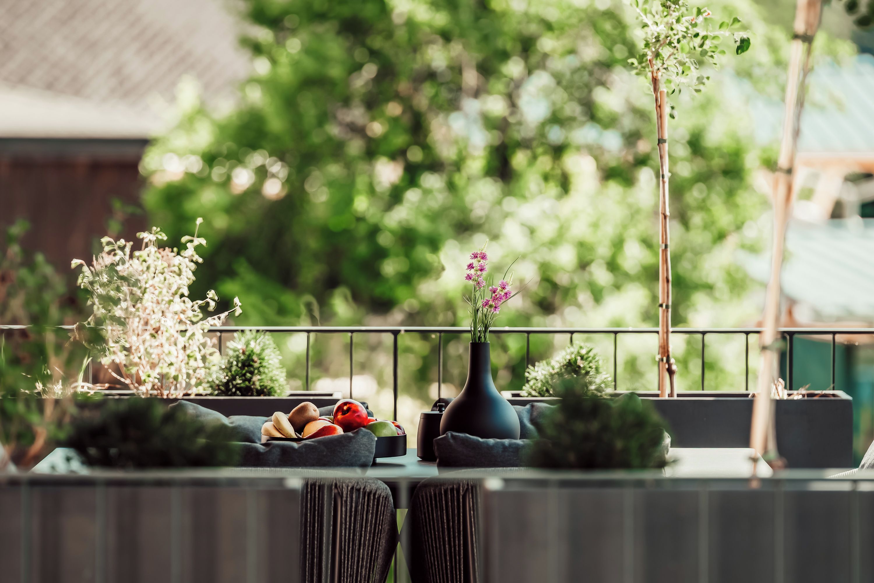 Ein Balkon mit Tisch, auf dem sich Obst und eine Vase mit Blumen befinden, umgeben von viel Grün.