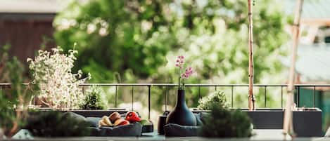 Ein Balkon mit Tisch, auf dem sich Obst und eine Vase mit Blumen befinden, umgeben von viel Grün.