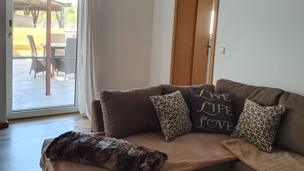 A living room with a brown sofa, patterned cushions, a glass coffee table, and a sliding glass door leading to an outdoor patio. - Holiday Home 'Island Pferdehof Arrisholt Solskin' with Private Terrace, Shared Garden & Wi-Fi (Kißlegg)