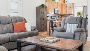 A living room with a sofa, armchair, and coffee table, a kitchen with wooden cabinets and appliances, and a view of the outdoors. - Modern Table Rock Lake Retreat | Swim Dock + Pool (Reeds Spring)