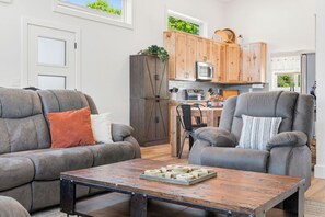 A living room with a sofa, armchair, and coffee table, a kitchen with wooden cabinets and appliances, and a view of the outdoors. - Modern Table Rock Lake Retreat | Swim Dock + Pool (Reeds Spring)