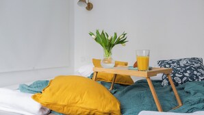 A bed with a teal blanket, a yellow pillow, and a white pillow. There is a small wooden table with a glass of orange juice and a vase with green leaves. - ShortStayPoland Orla B61 (Warsaw)