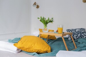 Ein Bett mit einer petrolfarbenen Decke, einem gelben Kissen und einem weißen Kissen. Ein kleiner Holztisch mit einem Glas Orangensaft und einer Vase mit grünen Blättern.