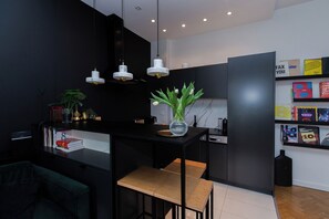 Eine moderne Küche mit schwarzen Schränken, einer zentralen Kochinsel mit einer Vase, Pendelleuchten und einem Bücherregal mit Büchern und Kartons.