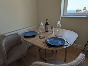 Ein Esstisch mit Tellern, Besteck, einer Weinflasche und Gläsern, in einem Raum mit Heizkörper, Stühlen und einem Fenster mit Blick.