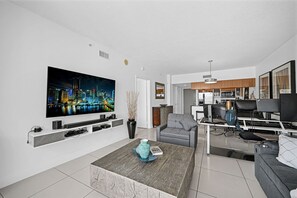 Ein modernes Wohnzimmer mit Flachbildfernseher, Couch, Couchtisch und Essbereich. Im Hintergrund ist eine Küche zu sehen.