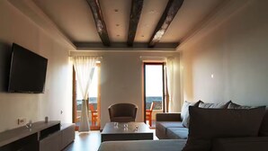 Un salon moderne avec un téléviseur à écran plat, un canapé, une table basse et un fauteuil. Il y a deux fenêtres avec des rideaux, et on aperçoit un balcon à travers l’une d’elles.