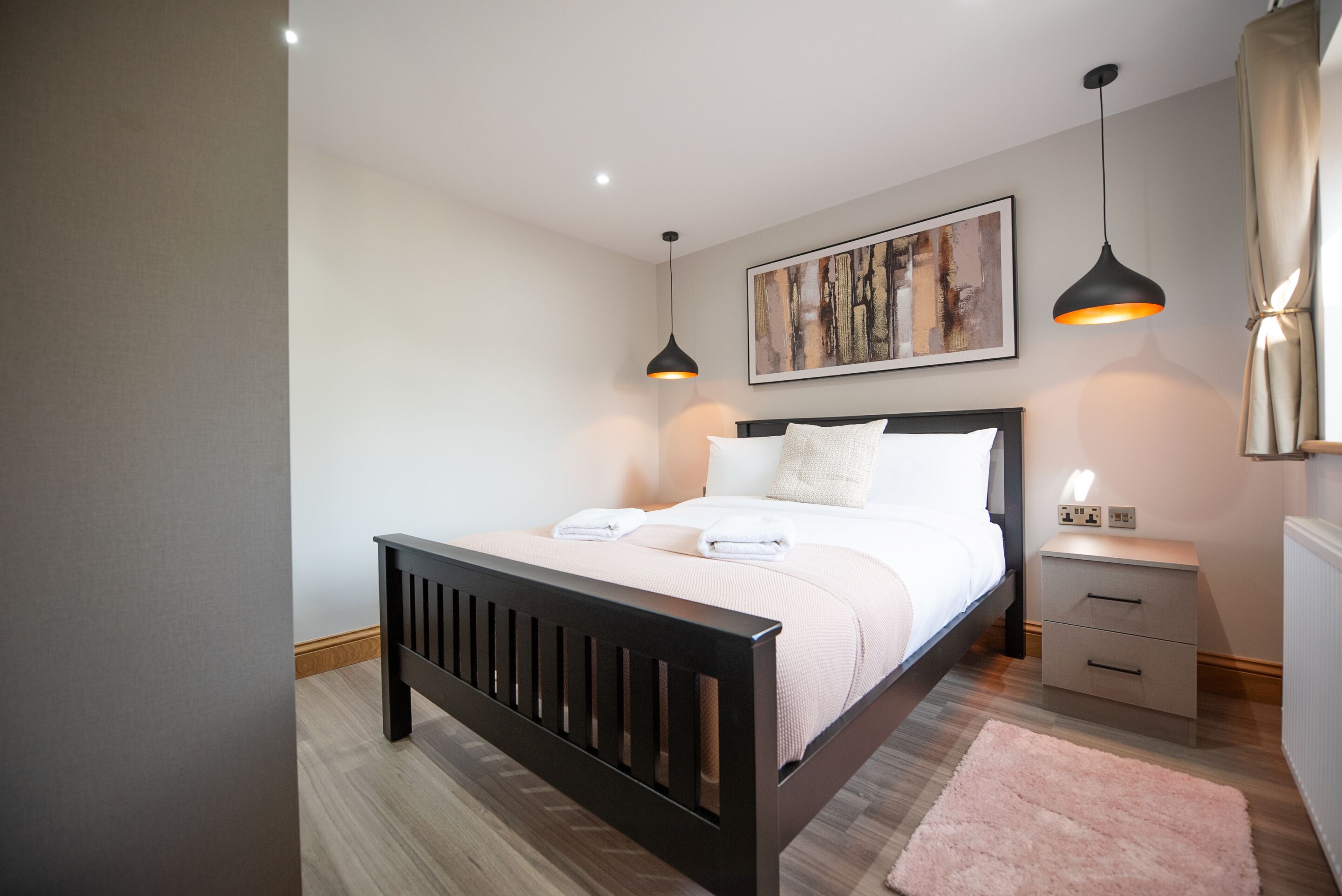 A bedroom with a bed, bedside tables, a nightstand, a hanging light, and a painting on the wall.