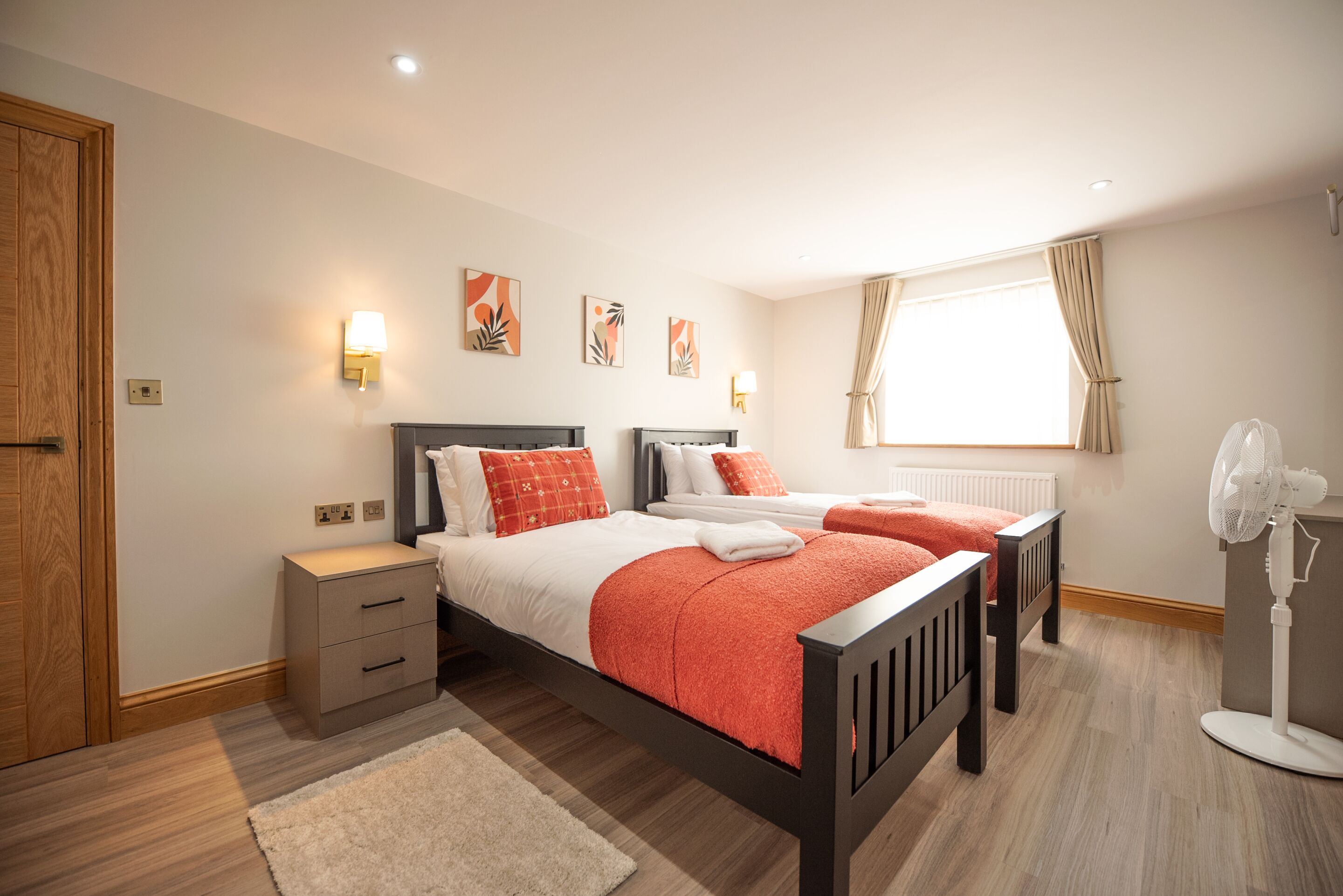 A bedroom with two beds, a nightstand, a bedside table, a standing fan, and a window with curtains.