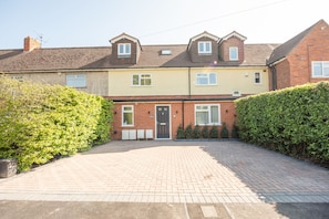 A two-story residential building with a paved driveway, a front door, and a small garden area.