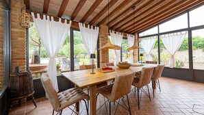 A covered dining area with a wooden table, cushioned chairs, and large windows.