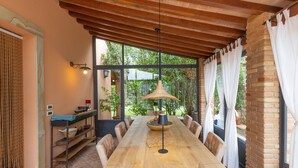 A covered dining area with a long wooden table, chairs, a hanging light fixture, and a view of greenery outside.