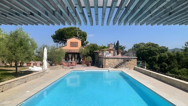 A swimming pool with a pergola overhead, a stone building with a chimney, and outdoor seating.