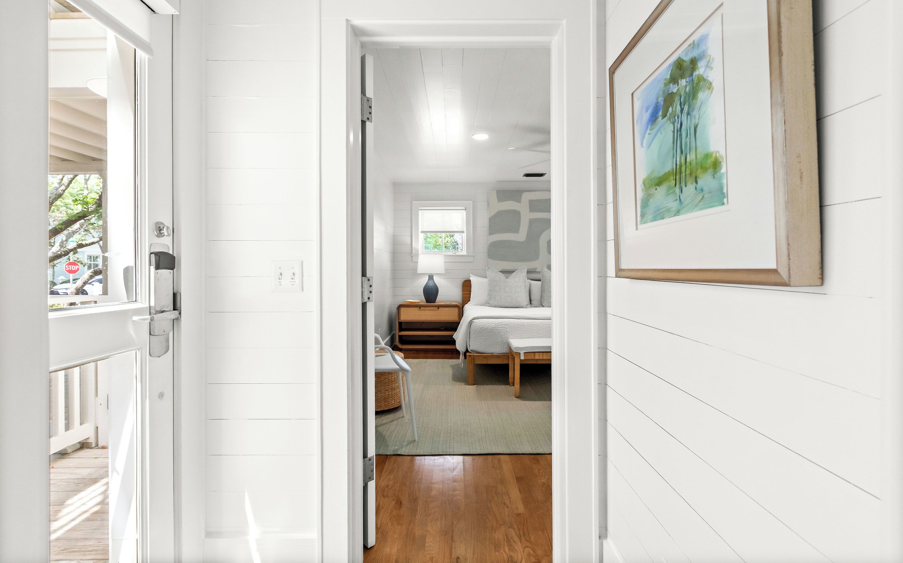 A hallway leading to a bedroom with a bed, bedside table, lamp, and a window.
